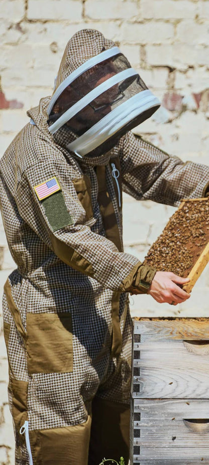 A person wearing a full beekeeping suit and face shield holds a wooden honeycomb frame covered with bees in front of a rustic brick wall.